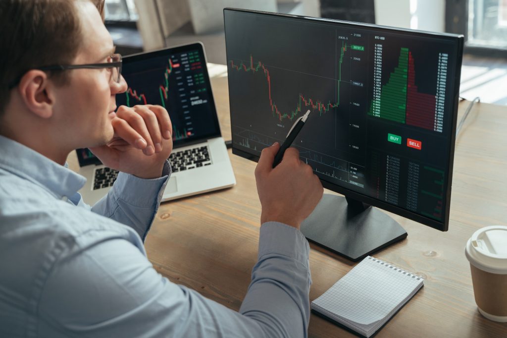 Concentrated man trader pointing pen at pc monitor with diagram of stock crypto currency market